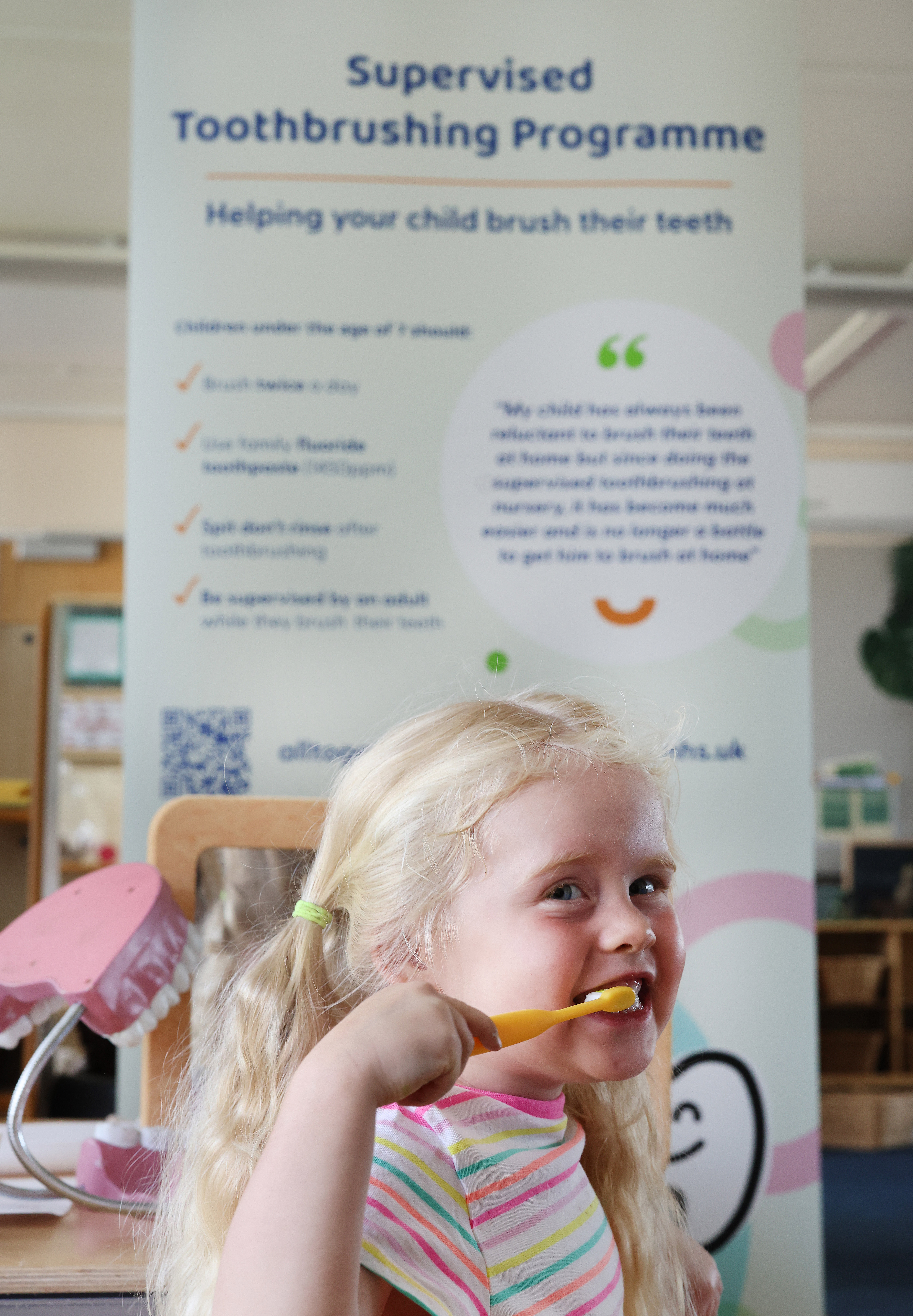 A smiling child with blonde hair brushes her teeth with a yellow toothbrush. Behind her, a poster titled "Supervised Toothbrushing Programme" offers guidance.