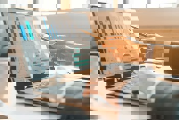 A person in business attire analyzes graphs on a laptop screen, pointing with a pen. Sunlight streams through a window.