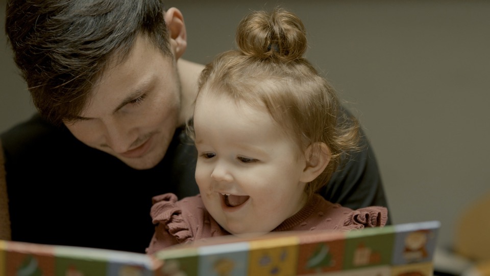 Father and young daughter share a joyful moment reading a colourful book. The child's face is lit up with excitement, conveying warmth and bonding.